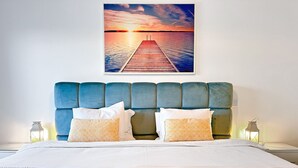 Un dormitorio con una cama grande, cabecera, dos mesitas de noche y un cuadro colgado en la pared con una vista de un atardecer sobre el agua.