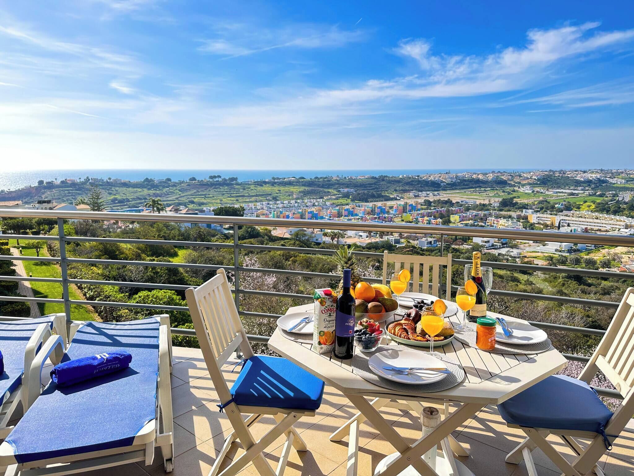 A dining table set with a bottle of wine, fruits, and glasses on a balcony overlooking a scenic landscape.
