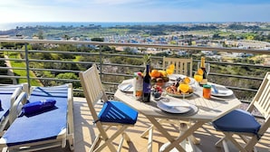 Una mesa de comedor dispuesta con una botella de vino, frutas y copas en un balcón con vistas a un paisaje pintoresco.