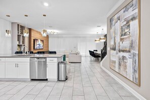 Una cocina moderna con armarios blancos, lavavajillas y un lavabo. Hay una zona de comedor con una mesa y sillas, y un cuadro abstracto grande en la pared.