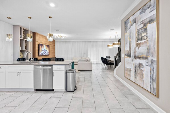 A modern kitchen with white cabinetry, a dishwasher, and a sink. There is a dining area with a table and chairs, and a large abstract painting on the wall. - 9BR house sleeps 24 with hot tub and pool (Kissimmee)