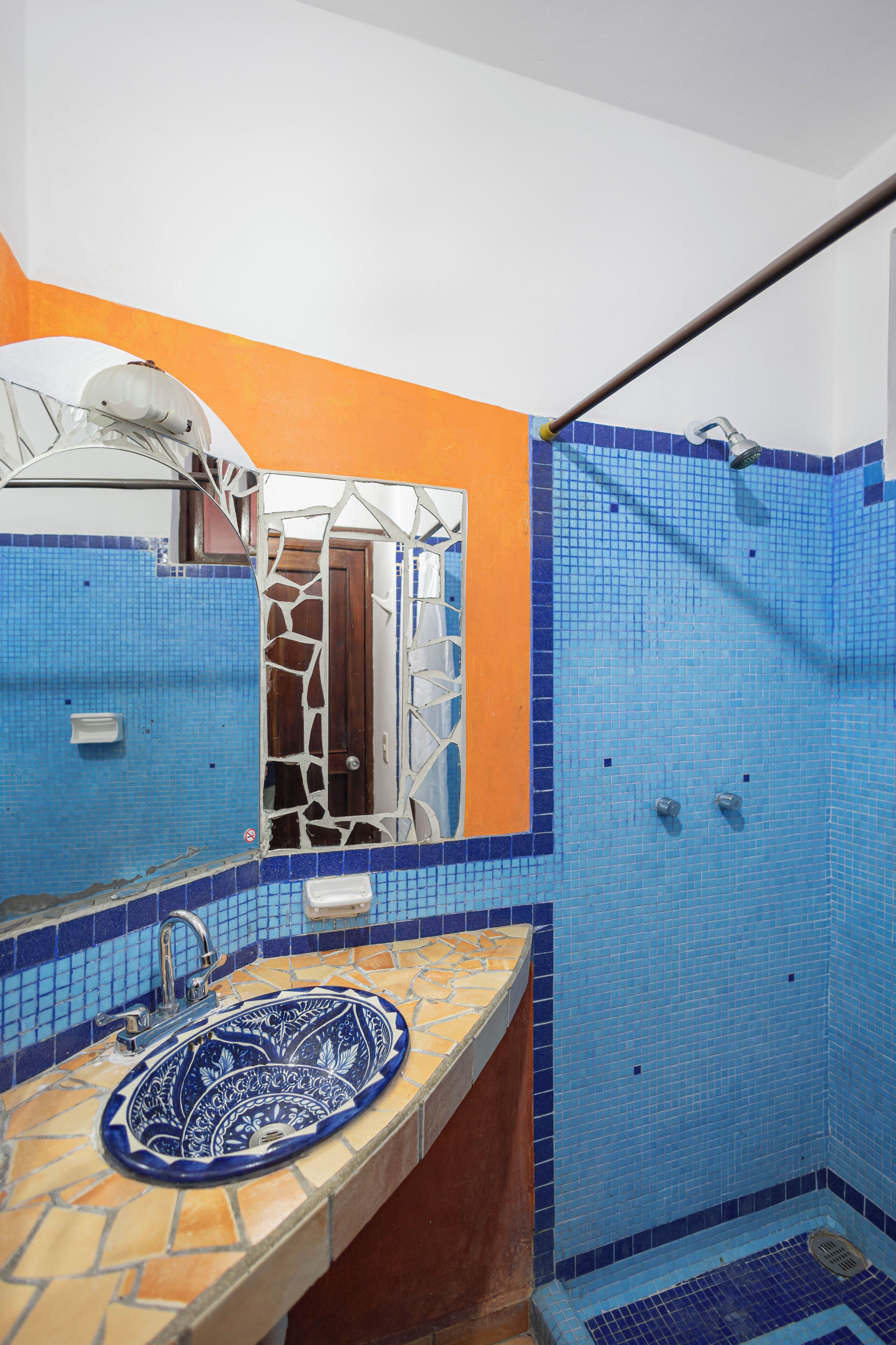 A bathroom with a blue-tiled shower, a sink with a decorative blue basin, and a mirror with a broken frame.
