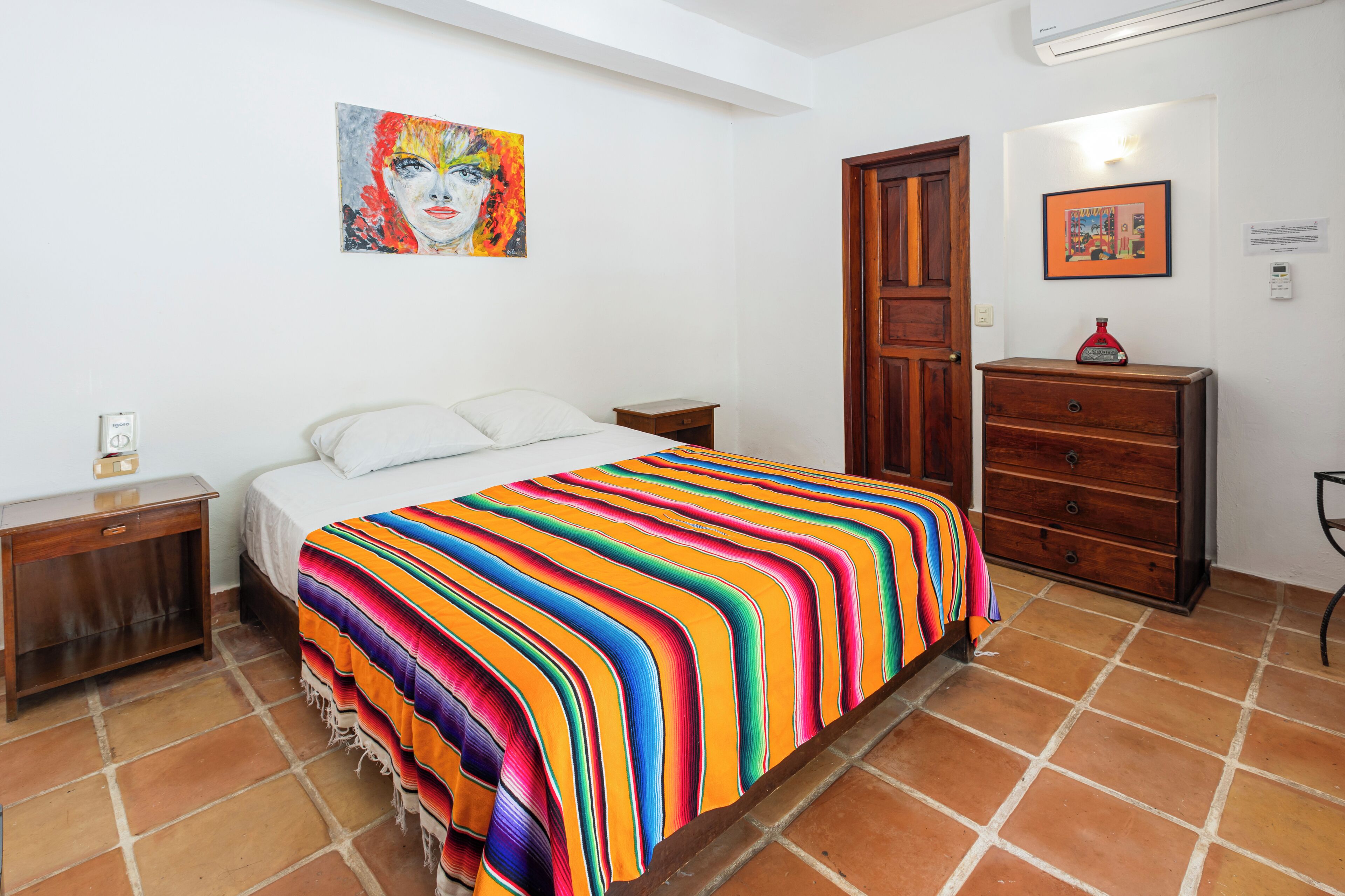 Un dormitorio con una colcha a rayas coloridas, un aparador de madera, una mesita de noche y un cuadro en la pared.