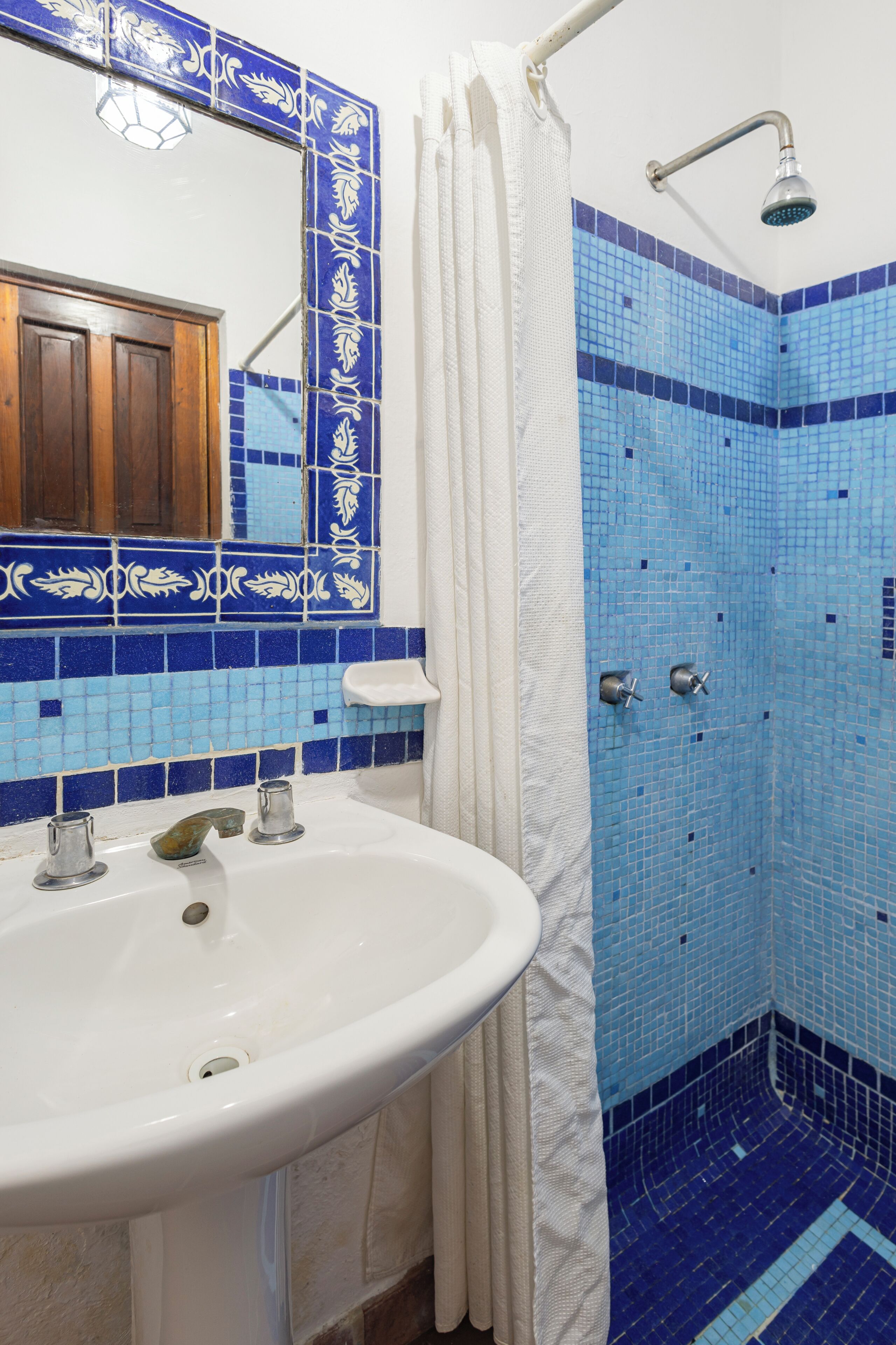 A bathroom with a white sink, blue mosaic tiles, a shower with a handheld showerhead, and a wooden door in the background.