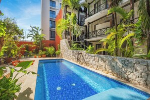 A swimming pool surrounded by lush greenery and stone walls, with a multi-story building in the background. - Casa Central  (Playa del Carmen)