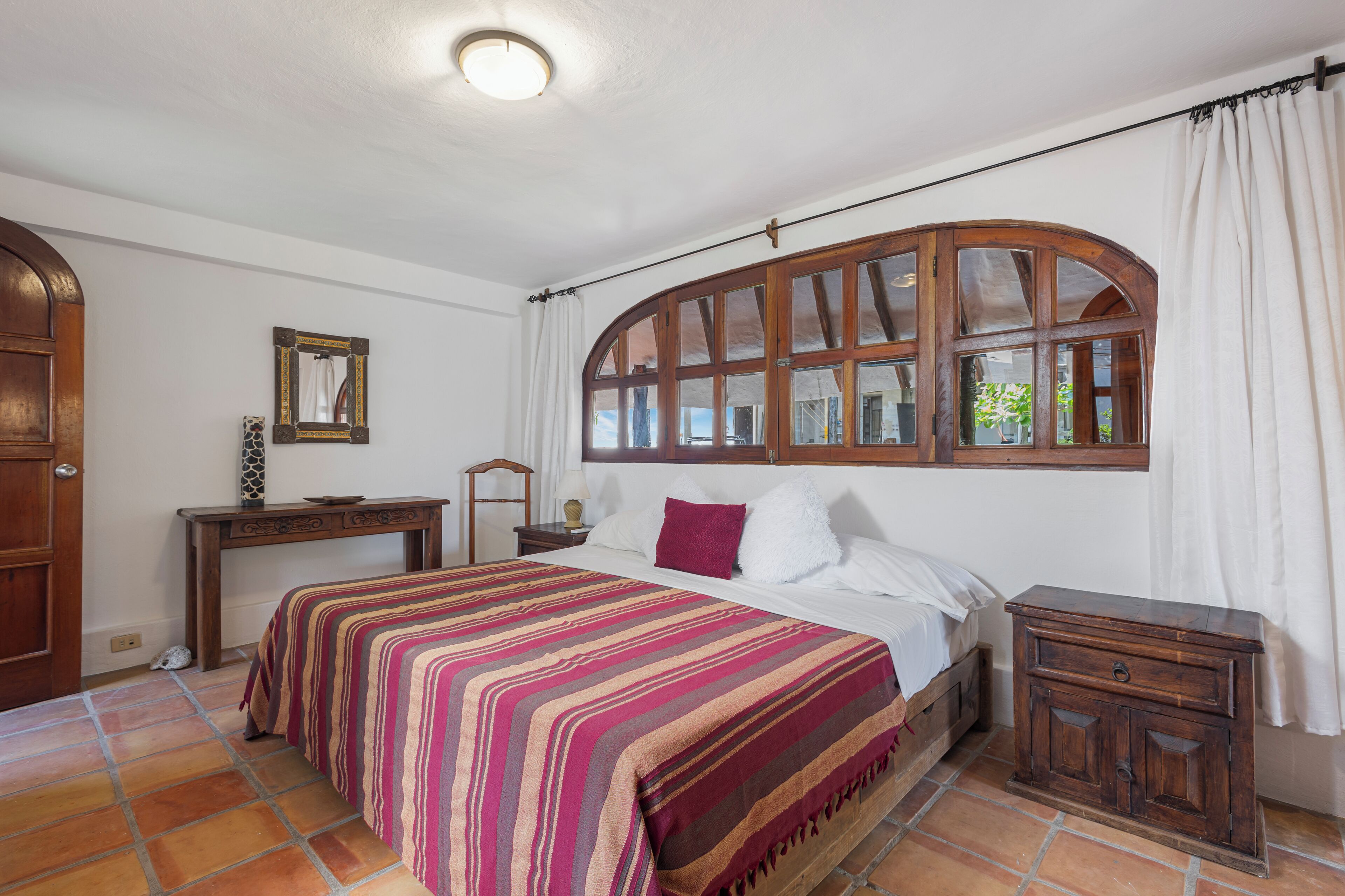 Un dormitorio con una cama grande, una mesita de noche de madera, un aparador, una ventana con cortinas y un suelo de baldosas.