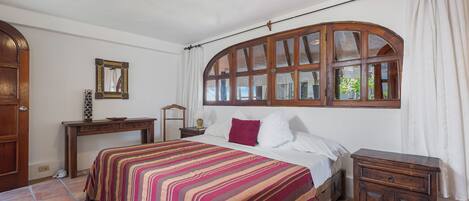Un dormitorio con una cama grande, una mesita de noche de madera, un aparador, una ventana con cortinas y un suelo de baldosas.