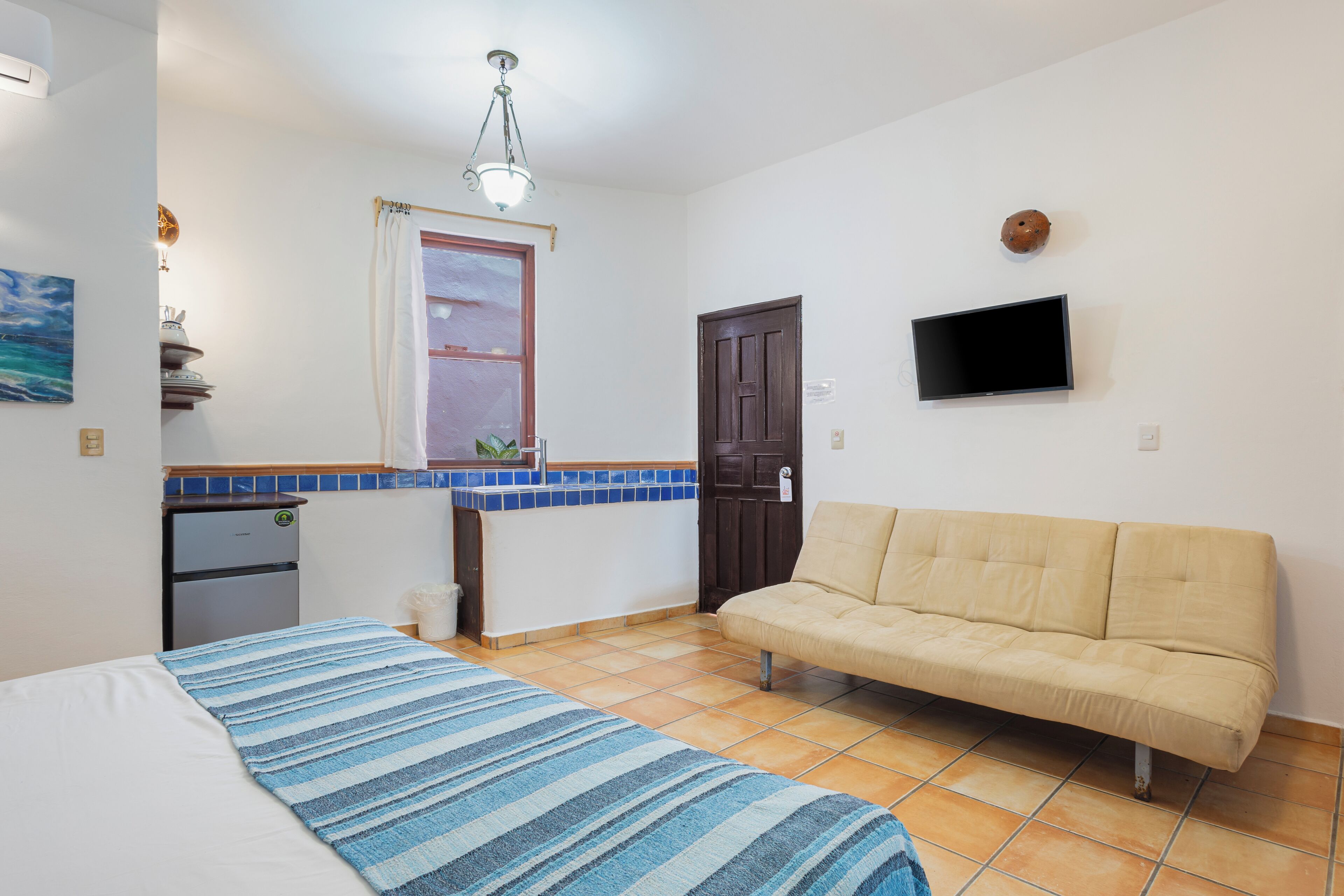 Quarto compacto com cama, sofá, pequeno espaço de cozinha e TV fixada na parede.