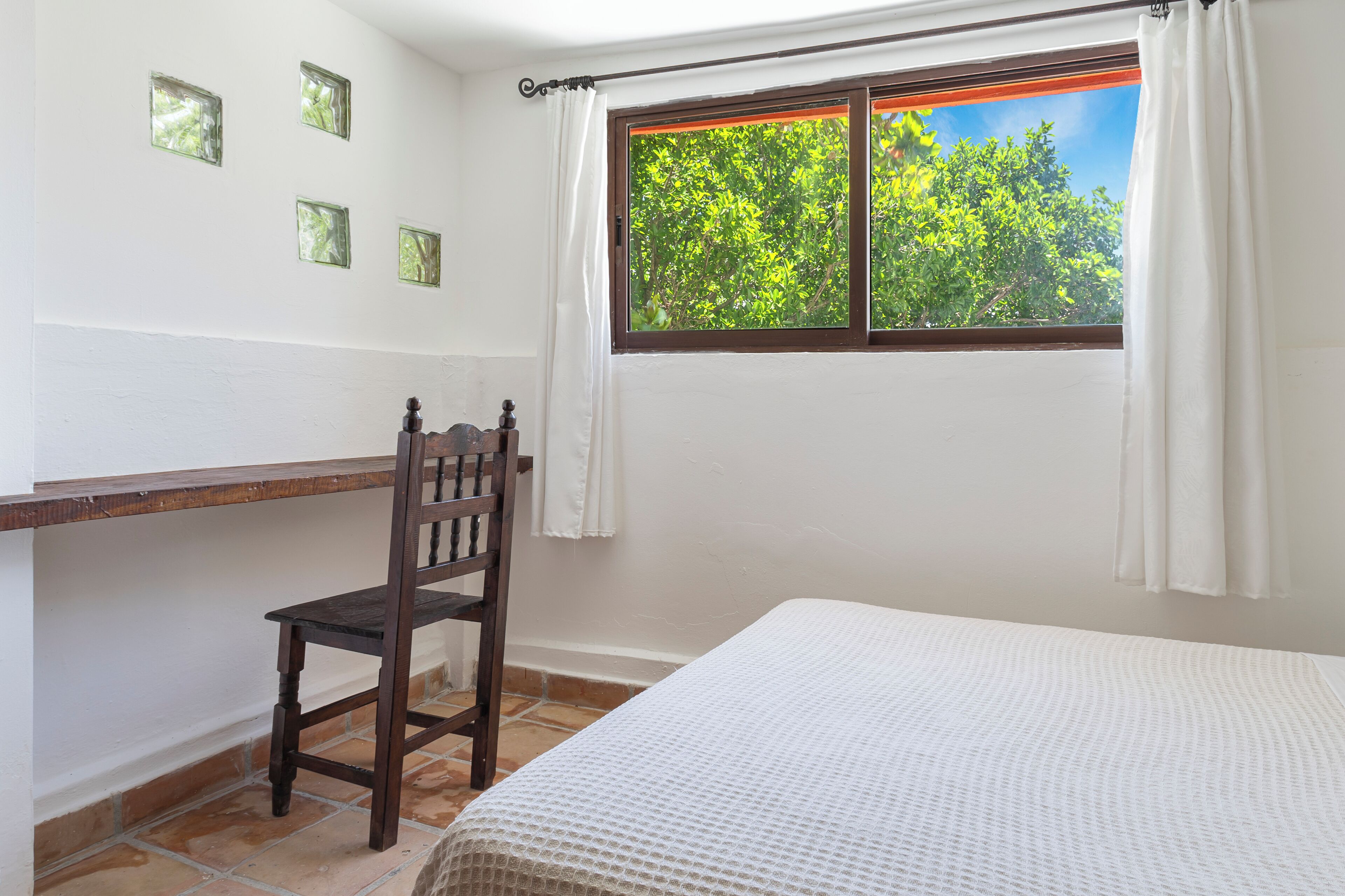 A bedroom with a bed, a wooden chair, a small table, and a window with curtains.