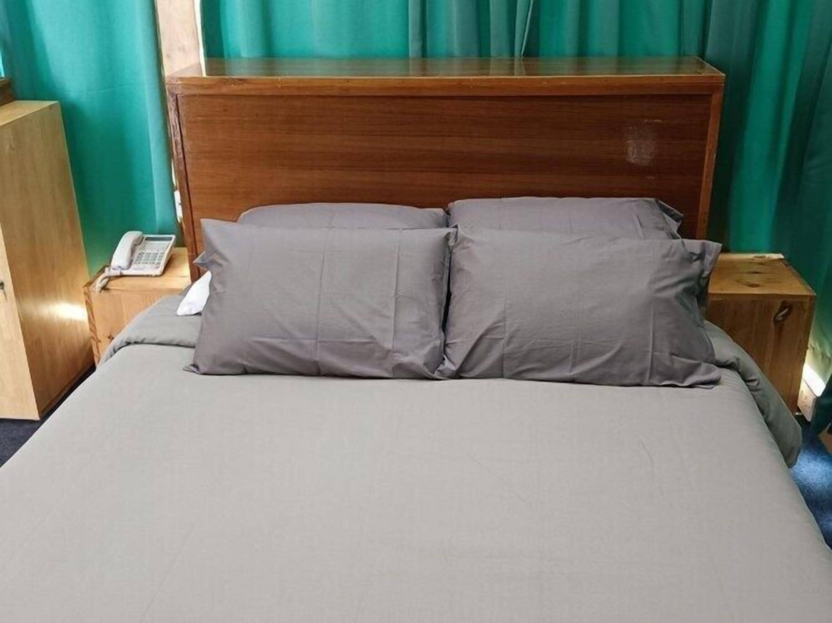 A bed with grey bedding and pillows, a wooden headboard, a bedside table with a phone, and green curtains.