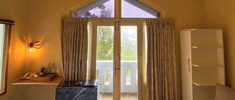 A hotel room with a bed, a desk with a computer, a wardrobe, and a view through a glass door to an outdoor balcony.