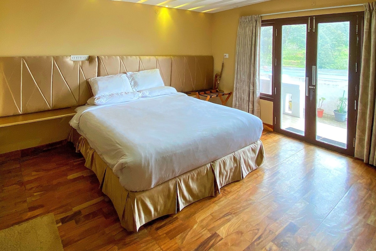 A spacious bedroom with a large bed, wooden flooring, and a view through the glass doors.