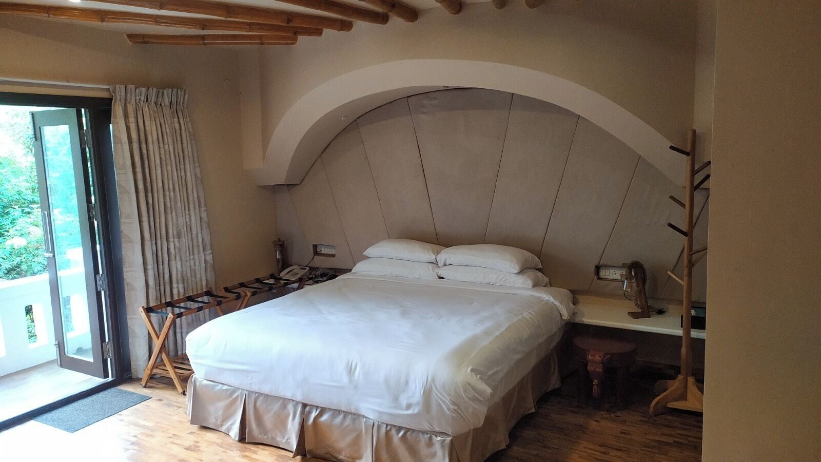 A bedroom with a large bed, a desk, and a chair. There is a window with curtains and a view of greenery outside.