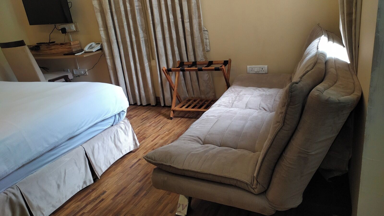 Een kamer met een bed, een slaapbank, een houten stoel, een bureau met telefoon en een aan de muur bevestigde televisie.