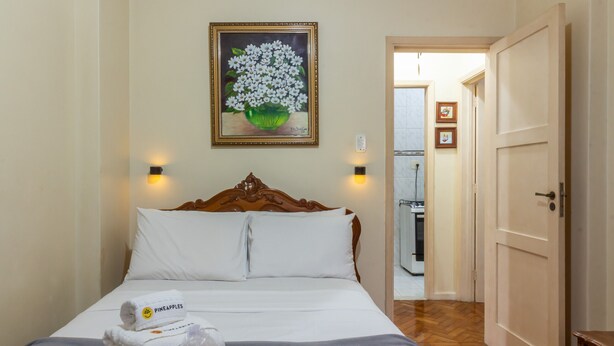 A neatly made bed with white linens and a Pineapple towels set, a painting of white flowers on the wall, and a wooden door to the right. - 5 Minutes From Copacabana Beach Bi307 (Rio de Janeiro)