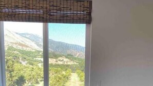 Ein Zimmer mit einem großen Fenster und Blick auf eine gebirgige Landschaft, einer Holzbank mit Kissen sowie einem gemusterten Fensterbaldachin.