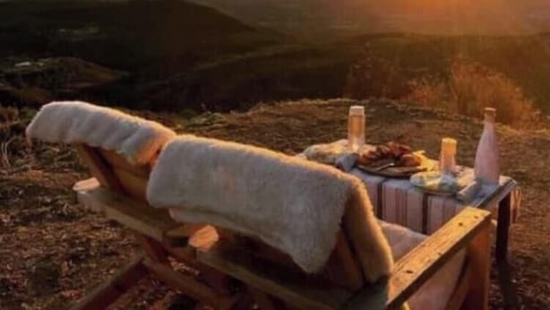 Ein hölzerner Liegestuhl mit Handtuch, Flasche und Glas auf einem Tisch, mit Blick auf eine Berglandschaft im Abendrot.