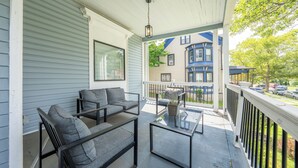 A covered porch with a sofa and a glass-top table, overlooking a street with parked cars and a blue building. - Casa Francesca 6BR Villa & Heated Pool (Roselle)