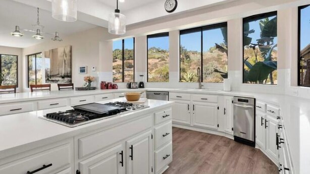 A modern kitchen with white cabinetry, a central island with a stove, and large windows offering views of the outdoors. - Hilltop Villa_Amazing Pool+Views (Fallbrook)