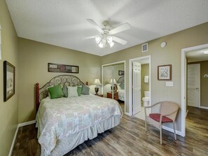 A bedroom with a bed, a chair, a ceiling fan, and a door leading to another room.