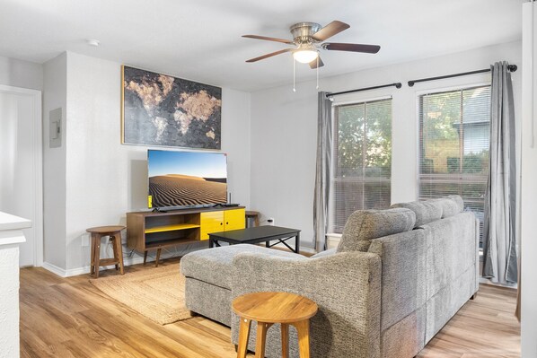 A living room with a grey sofa, a wooden coffee table, a TV on a yellow stand, and a ceiling fan. - Beautiful 1BD/1BA in University Hills 99tn (Austin)