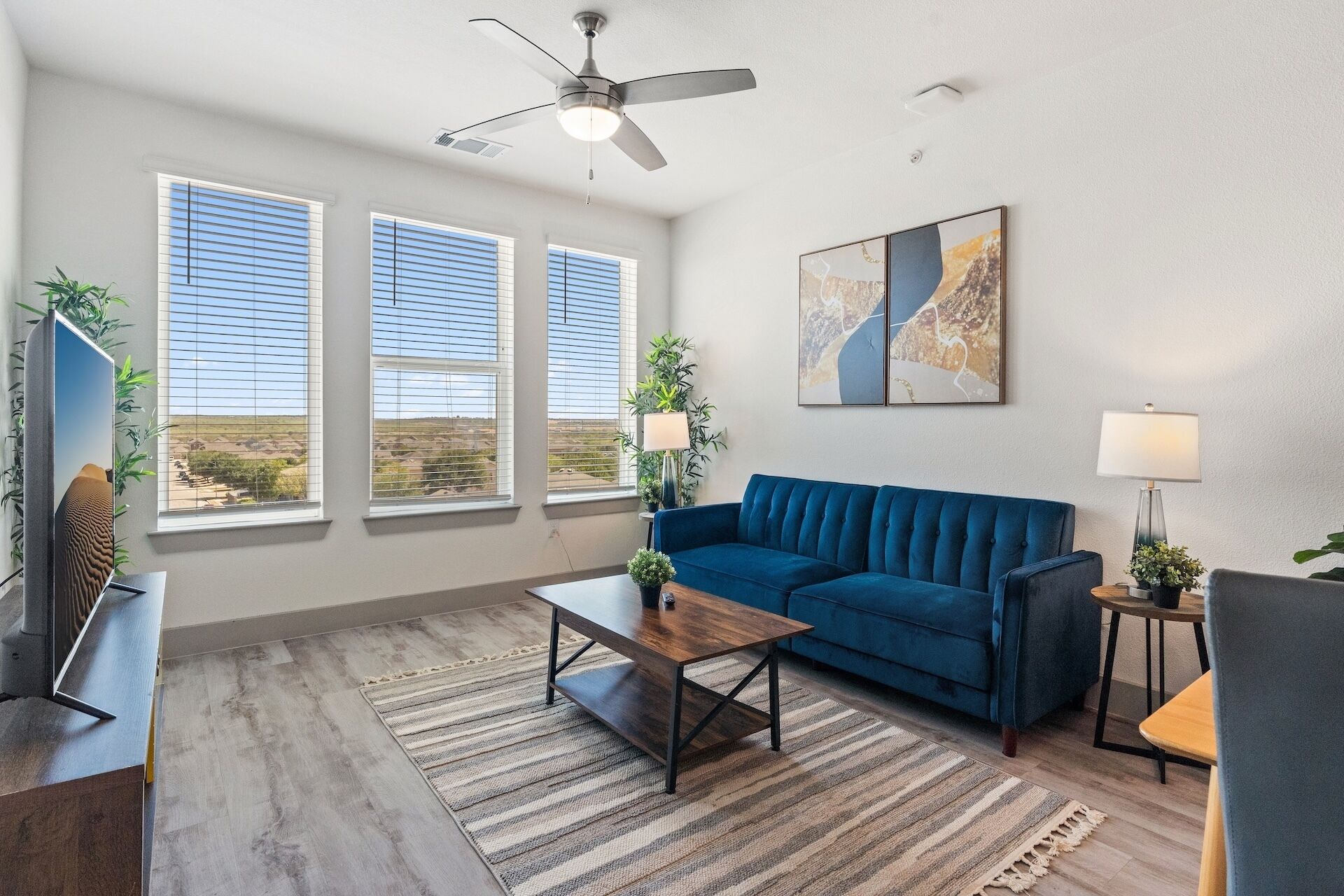 A modern living room with a blue sofa, a wooden coffee table, and a ceiling fan. There are three windows with blinds, a painting on the wall, and a lamp on a side table.