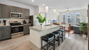 A modern kitchen with stainless steel appliances, a central island with bar stools, and a dining area with a view of the city. - Luxury 2BD/2BA Next to Airport fyo2 (Del Valle)