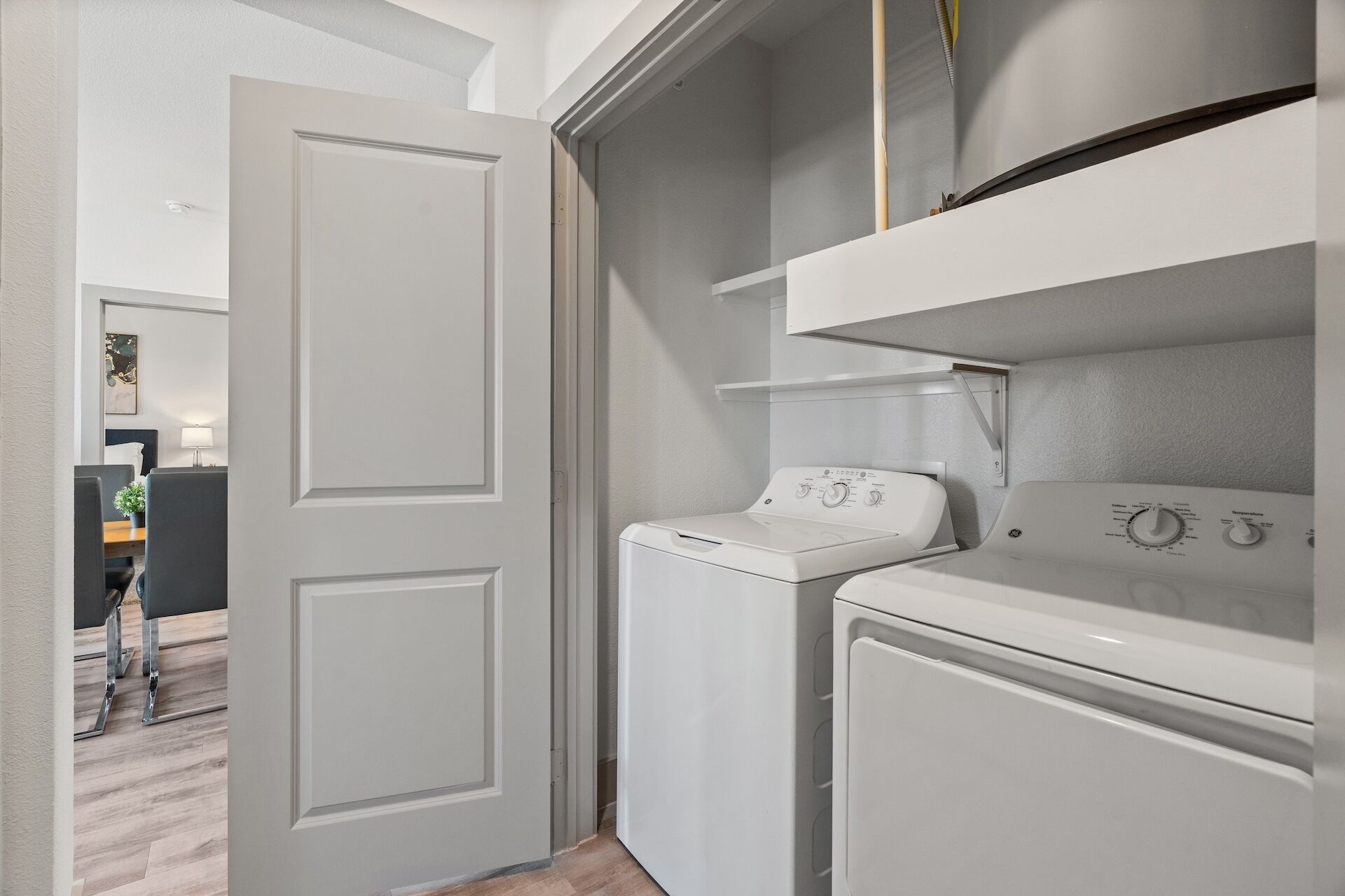 A laundry room with a front-loading washer and dryer, a small shelf above the dryer, and a door leading to another room.
