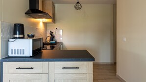 A compact kitchen with a microwave, kettle, and coffee maker on the counter, wooden cabinets, and a hanging light fixture. - Le Reflet de l'Horizon - Proche Plage (Saint-Quentin-la-Motte-Croix-au-Bailly)