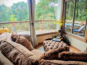 A cozy room with a sofa and ottoman, a Buddha statue, and a view of a wooden deck and greenery outside. - Mountain Retreat • Sauna • Hot Tub • Healing & Meditation • Scenic Views (Waynesville)