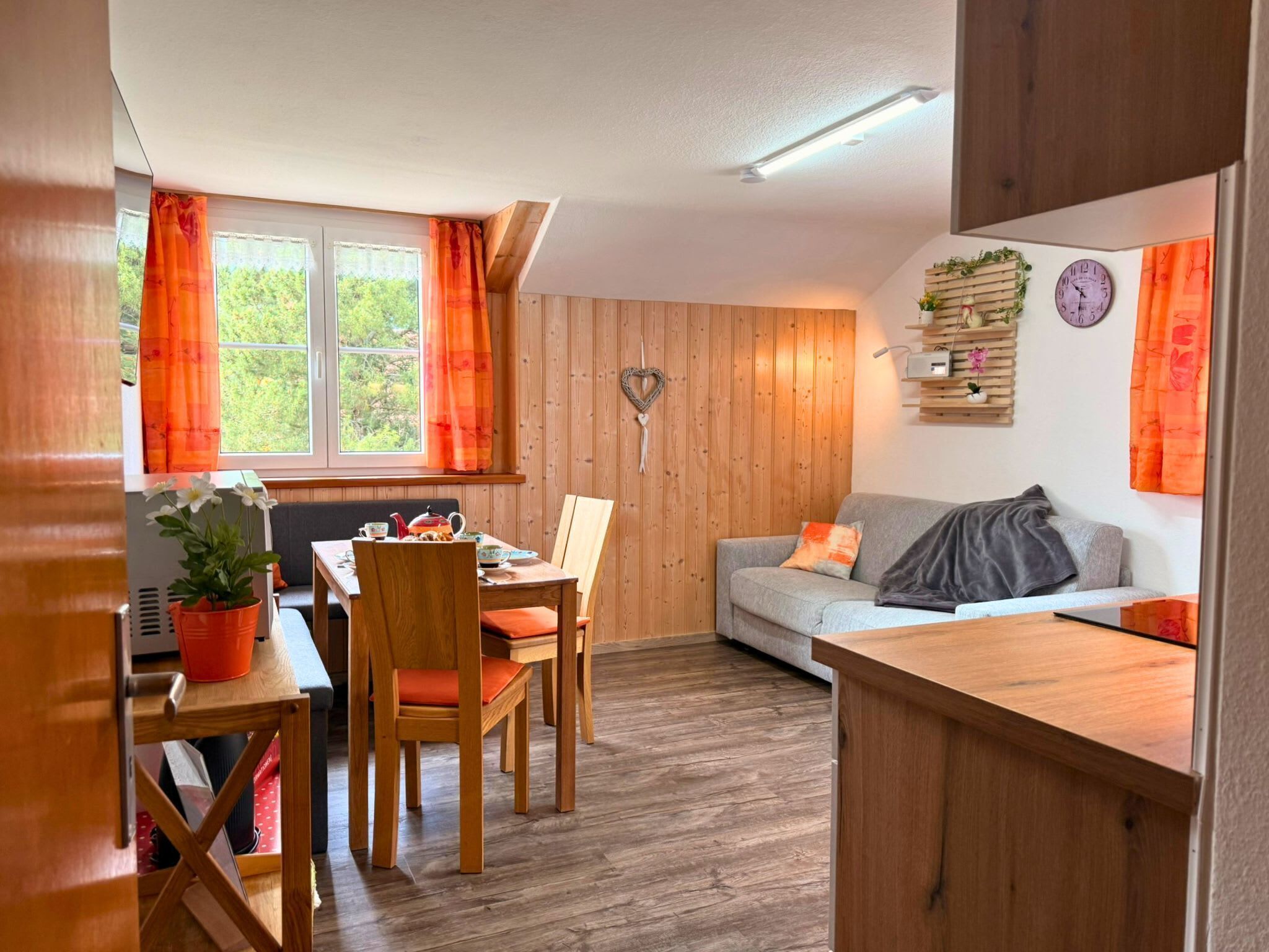 Ein gemütliches Zimmer mit Holzboden, einem Esstisch mit Stühlen, einer Couch und einem Fenster mit Vorhängen.