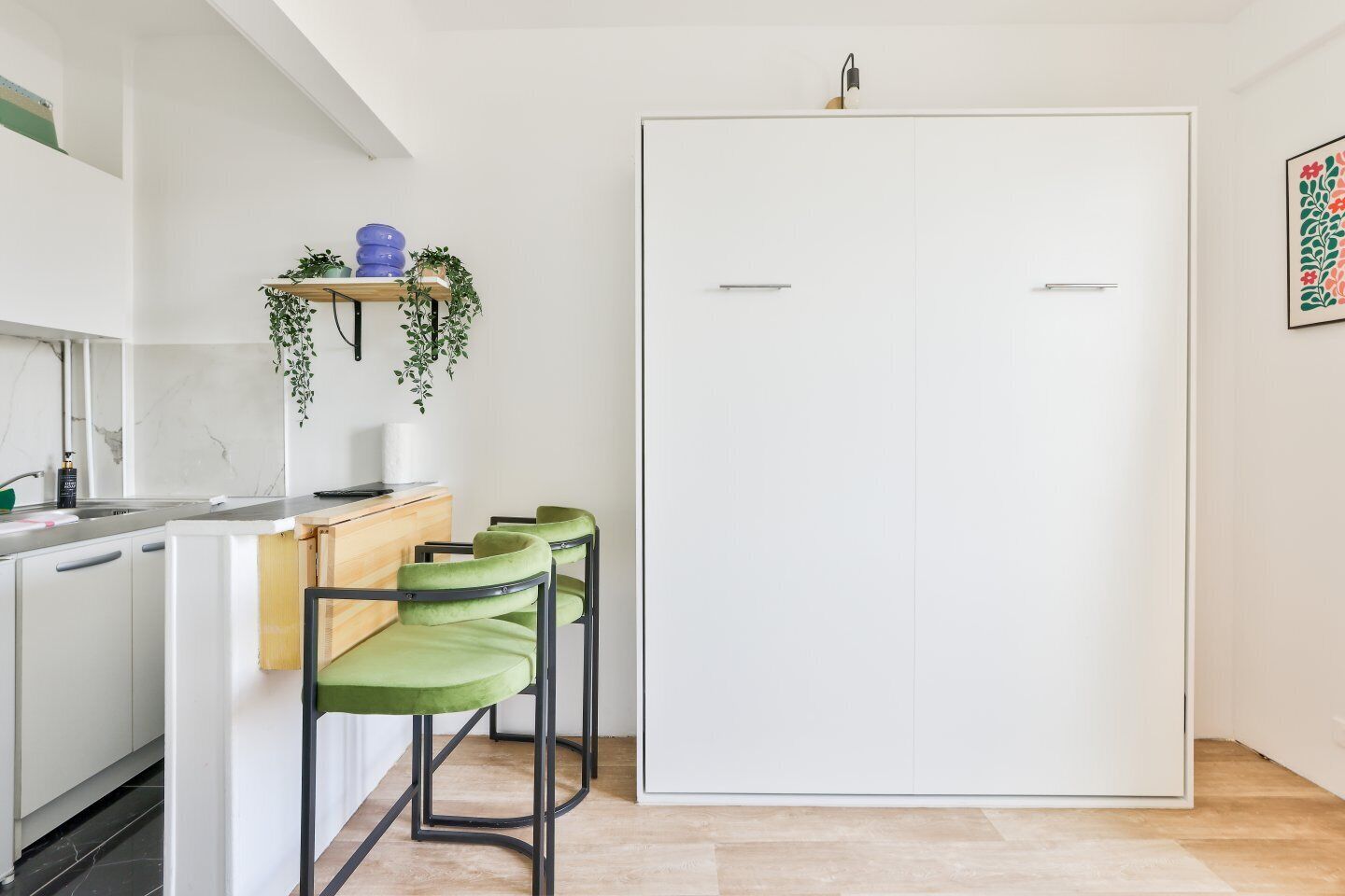 Een compacte keuken met een witte deur, een klein barretje met groene stoelen en een plank met planten en een blauwe container.