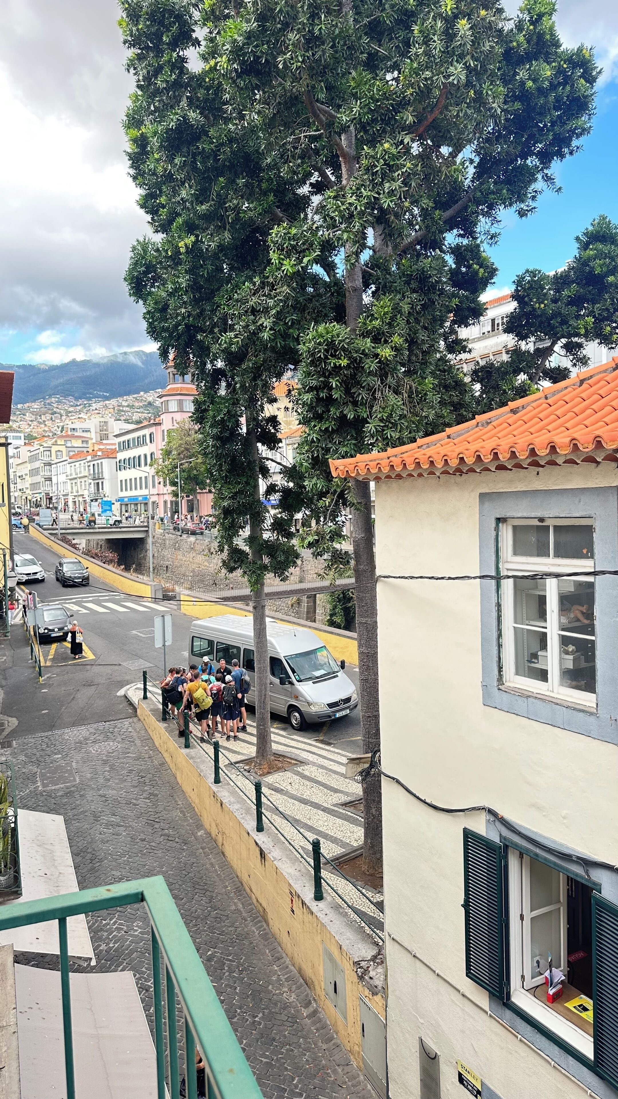 Una veduta di strada con un albero, un edificio con tetto in tegole e un gruppo di persone in piedi sul marciapiede.