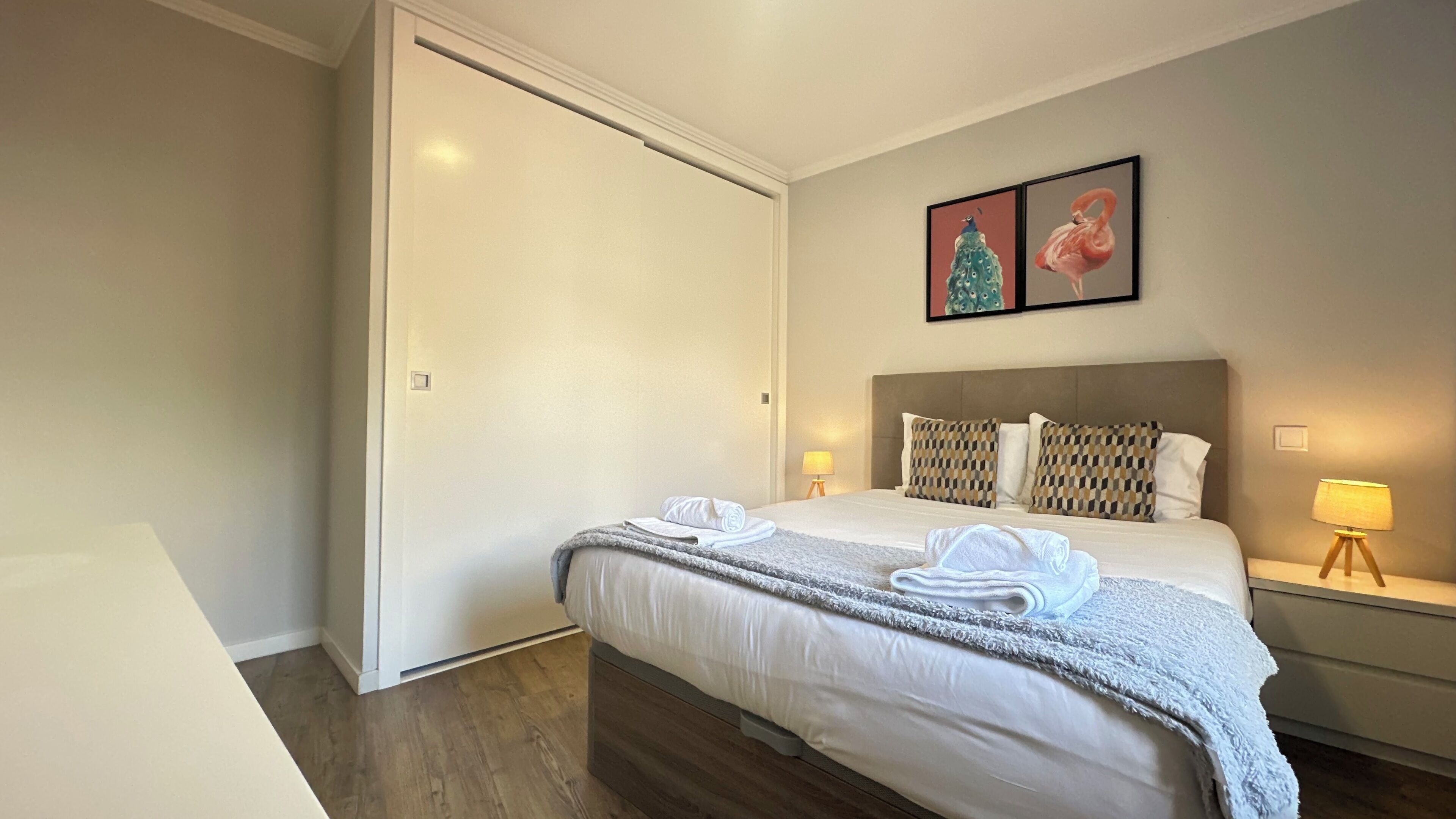 Habitación de hotel con cama, armario, mesita de noche con lámpara y dos cuadros colgados en la pared.
