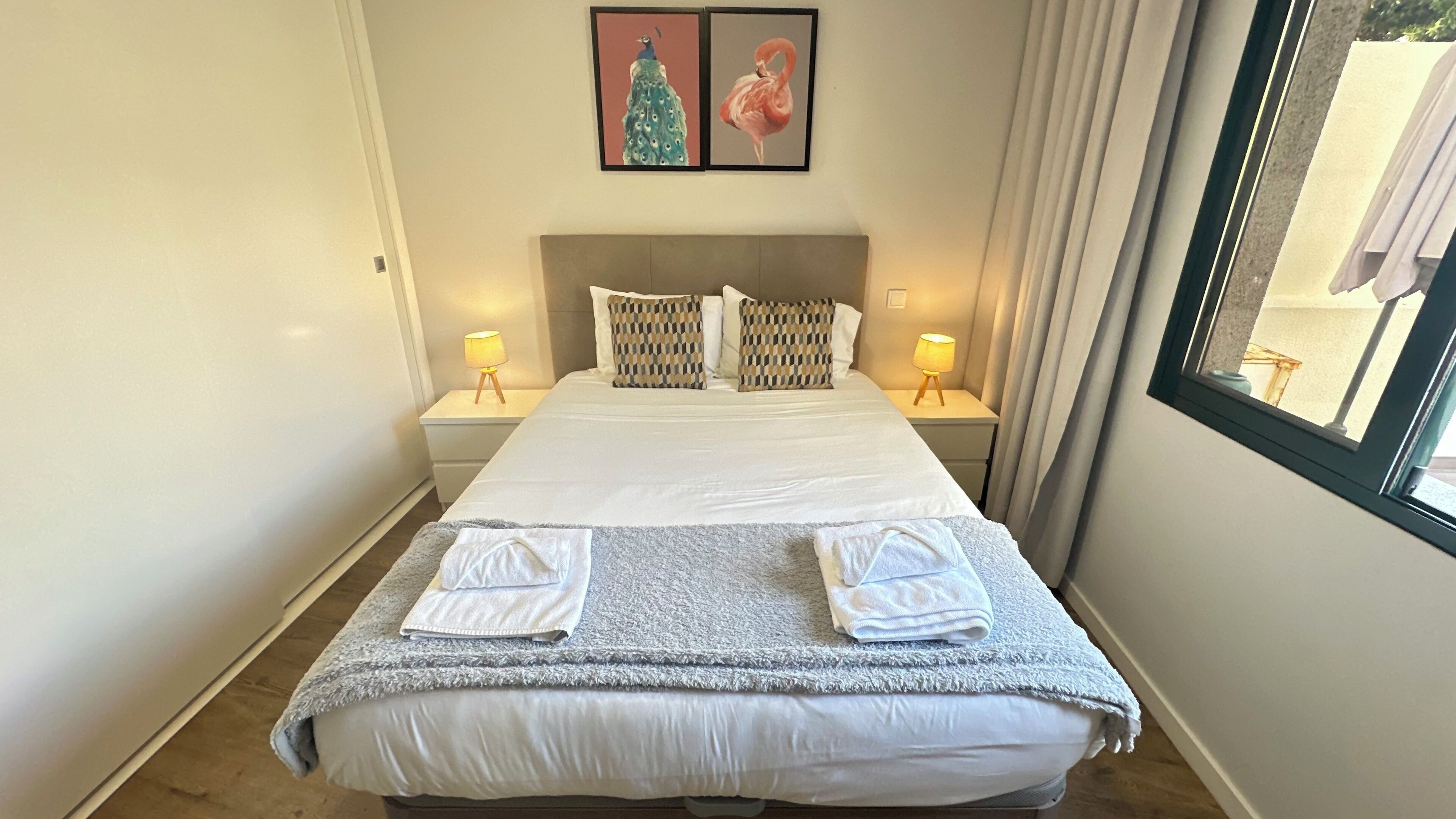 A neatly made bed with two pillows, a grey blanket, and two towels. There are two bedside lamps on either side of the bed.
