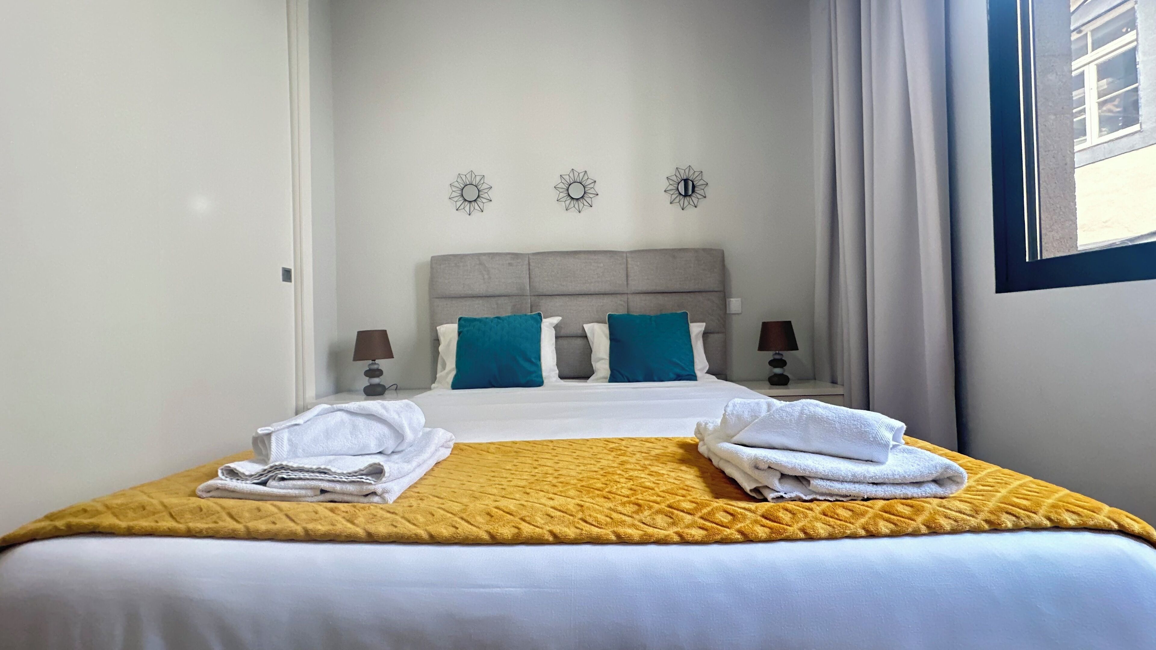 A neatly made bed with white linens and two towels, a grey headboard, and two teal pillows.