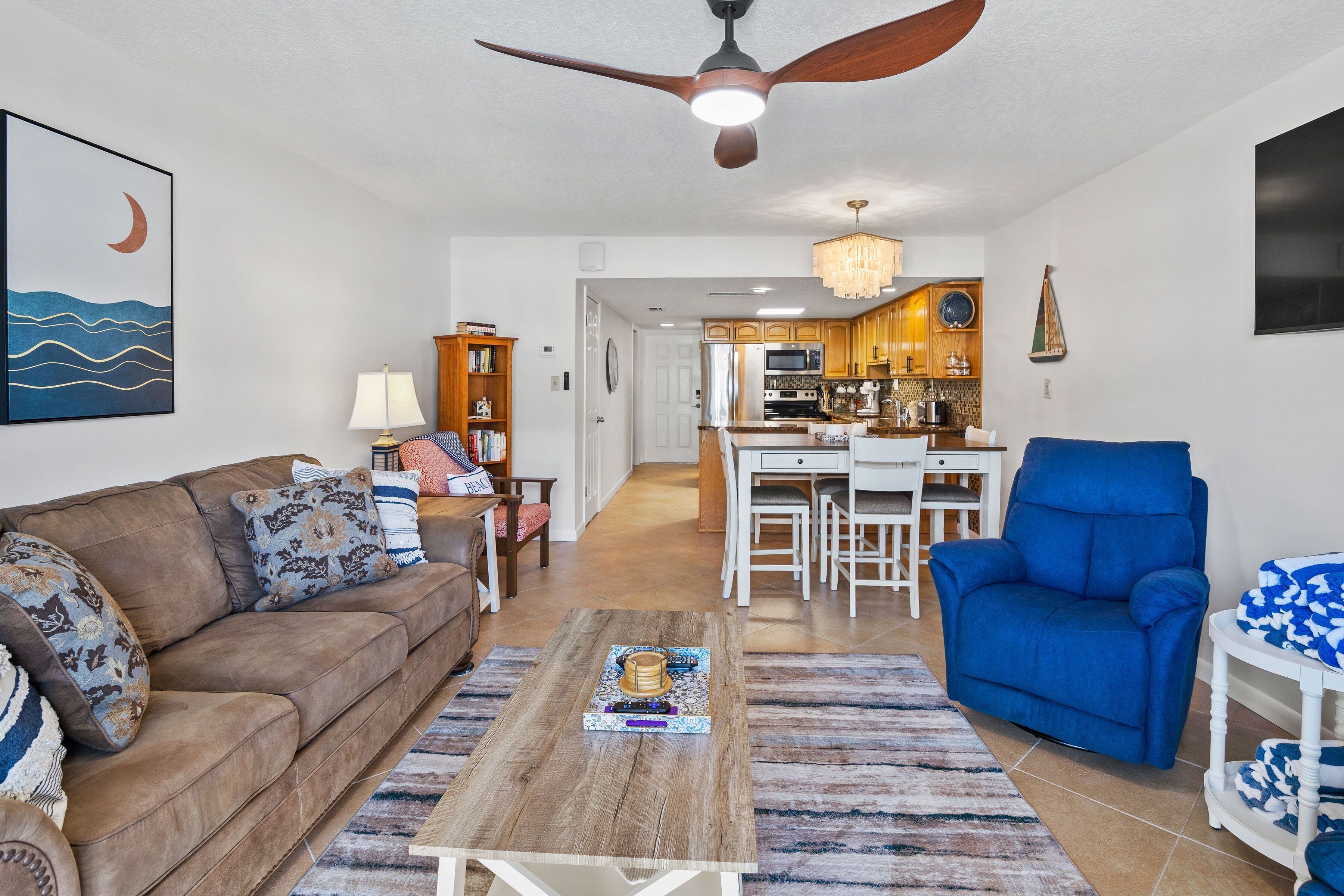 A living room with a brown sofa, a blue armchair, and a coffee table. There is a kitchen in the background with wooden cabinets and a chandelier.