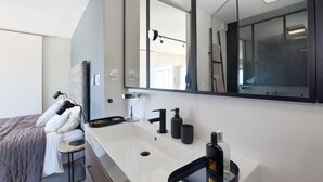 Un baño moderno con un espejo grande, un lavamanos blanco y un grifo negro. Hay un perchero con una toalla y una silla negra.