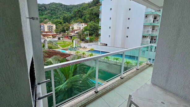 A balcony with a view of a swimming pool, a building with balconies, and a green hillside.