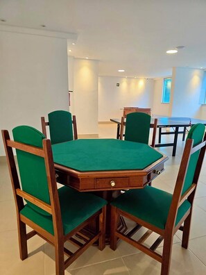 Un salón de conferencias con una mesa rectangular y seis sillas, cada una con asiento y respaldo tapizados en verde. La sala tiene paredes blancas, iluminación empotrada en el techo y un ventanal con persianas.