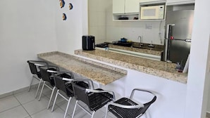 A compact kitchen with a granite countertop, white cabinets, a microwave, and a stainless steel refrigerator. There are four black chairs with metal legs.