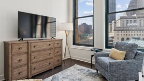 Een moderne woonkamer met een groot raam, een flatscreen televisie, een houten dressoir, een grijze bank en een vloerlamp.