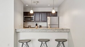 A modern kitchen with dark cabinets, stainless steel appliances, and a granite countertop. There are three bar stools under the counter. - Downtown Pittsburgh | Steps to Convention Center & Dining | Kitchen & Gym (Pittsburgh)