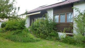 A house with a sloped roof, a wooden front porch, and a window with a curtain. - Holiday Home "Unteres Hart" – Spacious and Comfortable (Gomadingen)