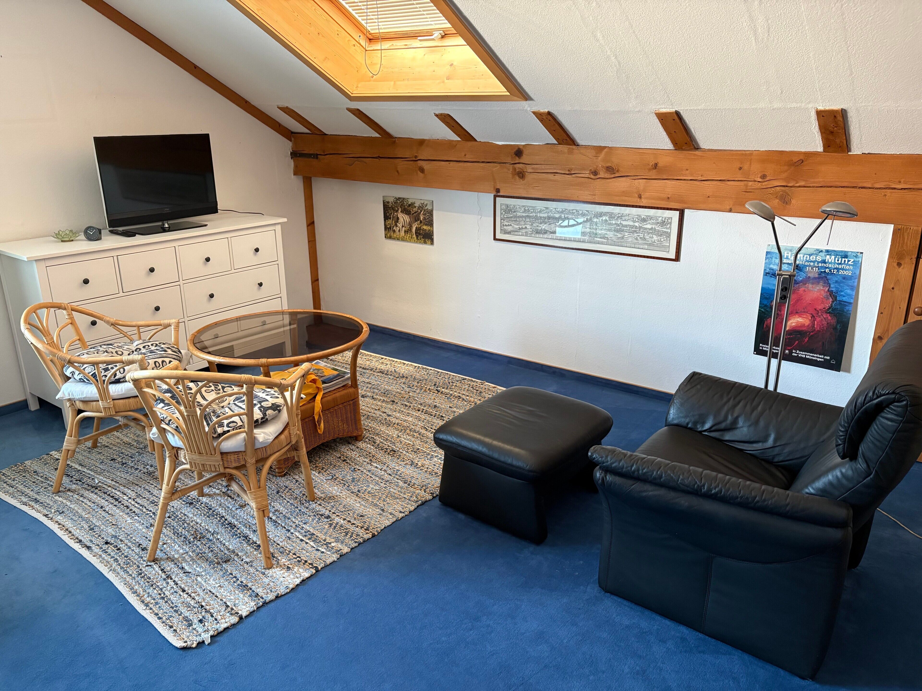 Ein Zimmer mit Fernseher, Korbmöbeln, einem Glastisch, einem schwarzen Ledersofa und einem blauen Teppich.