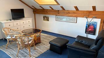 Ein Zimmer mit Fernseher, Korbmöbeln, einem Glastisch, einem schwarzen Ledersofa und einem blauen Teppich.
