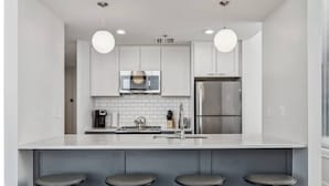 A modern kitchen with white cabinets, a stainless steel refrigerator, a microwave, and a coffee maker. There are three stools under the counter.