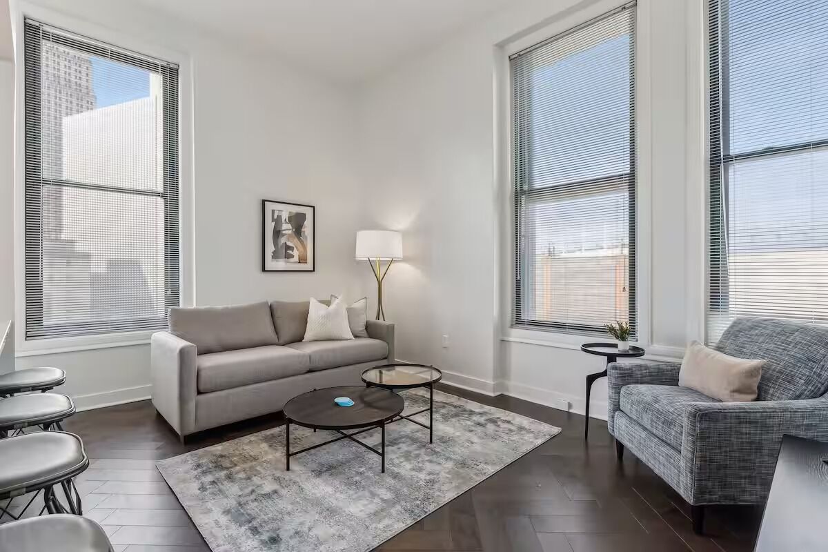 Un salon moderne avec un canapé gris, une table basse ronde et un fauteuil gris. On y trouve deux petites tables d’appoint, l’une avec une lampe et l’autre avec une petite plante.