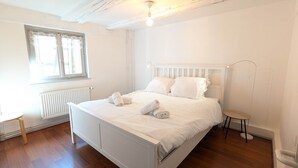 Un dormitori blanc amb un llit, un radiador, una finestra amb cortines i una taula petita.