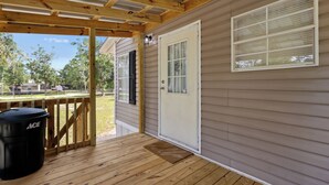 Eine überdachte Veranda mit Holzdeck, eine weiße Tür mit Fenster und ein schwarzer Mülleimer mit der Aufschrift 'ACE'.